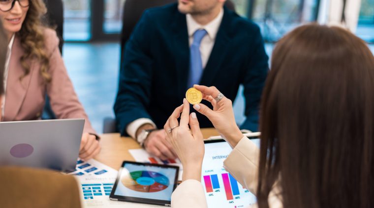 Female worker holding a physical cryptocurrency coin in an office. Gadgets and papers on the table